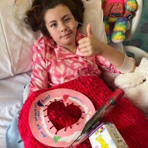 Grace sits up in her hospital bed giving a thumbs up, wearing pink heart-patterned pyjamas and with a nasal feeding tube. On her lap is a handmade Valentine's card made from a paper plate with a red woven yarn heart, reading 'Happy Valentines, get well soon Grace, from DK'. A rainbow teddy bear sits beside her pillow, and a red rose and box of chocolates rest on her red blanket.