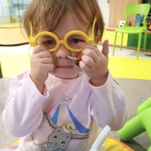 Toddler playing with oversized yellow toy glasses in a hospital playroom.