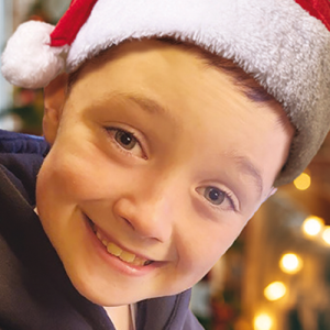 A close-up of George, a smiling young boy wearing a red Santa hat, with warm Christmas lights and a decorated tree glowing in the background