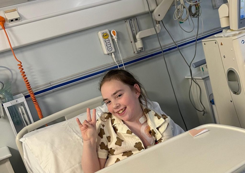 A smiling girl in a hospital bed giving a peace sign