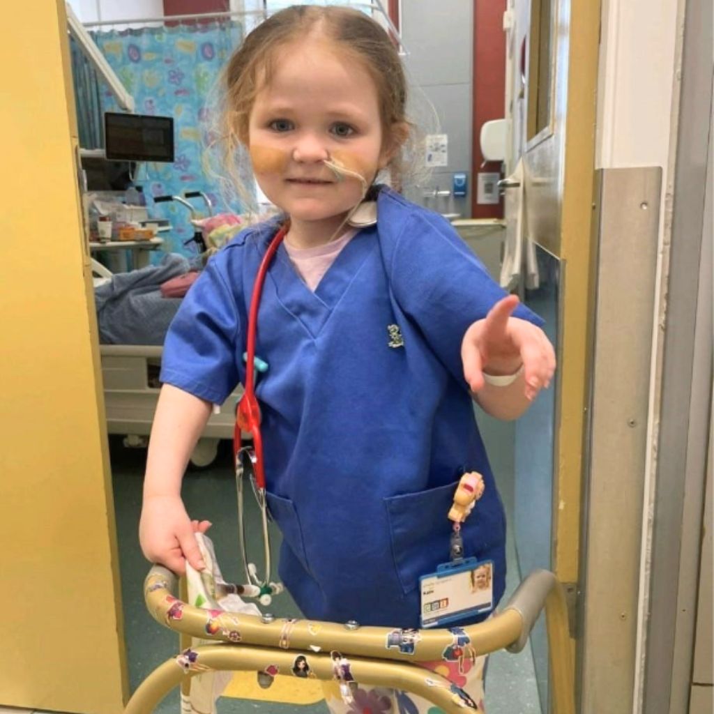 Katie dressed up as a nurse in scrubs with a stethoscope and ID badge, standing with her sticker-covered walking frame.